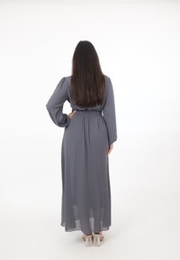 Mujer con cabello largo y castaño de espaldas, vestida con un vestido largo gris y sandalias con tacón transparente sobre un fondo blanco.