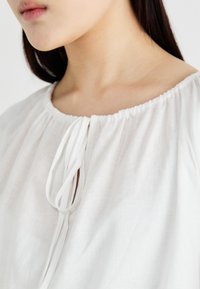 Blouse en lin blanc avec un col élastiqué et des détails de cordon. Texture lisse, coupe ample et drapé subtil.