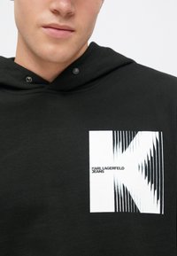 Felpa con cappuccio nera in cotone, caratterizzata da una grande grafica bianca della lettera "K" con un design a strisce e il testo "KARL LAGERFELD JEANS."