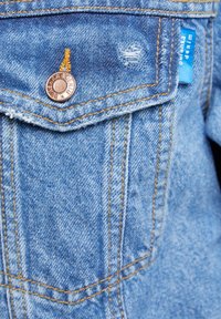 Light blue denim jacket pocket featuring a copper button, frayed edges, yellow stitching, and a small blue label. Includes a distressed detail.