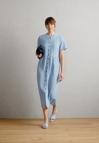 Vestito azzurro chiaro a maniche corte con bottoni frontali, vita arricciata e motivo a pois. Indossato con sandali azzurri chiari e una borsa nera.