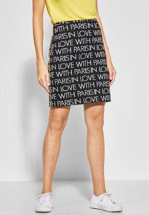 Kobieta w czarnej spódnicy z powtarzającym się białym napisem "IN LOVE WITH PARIS", połączona z żółtą bluzką i białymi trampkami.