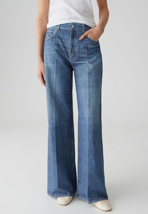 Persoon draagt een hoge taille, rechte blauwe jeans met voorvouwen en witte puntige schoenen, staand tegen een effen achtergrond.