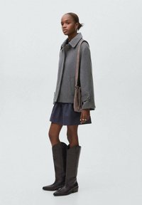 Femme portant un manteau en laine gris, une jupe bleu marine, des bottes noires à mi-mollet et portant un sac à épaule taupe, se tenant devant un fond clair uni.