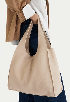 Persona con chaqueta beige y camisa blanca sosteniendo un gran bolso tote de cuero beige con textura, con asas anchas y pantalones vaqueros azules.