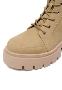 Beżowe zamszowe botki za kostkę z wiązaniem, okrągłym noskiem i grubą beżową podeszwą z rowkami zwiększającymi przyczepność. Srebrne oczka dodają szczegółu.