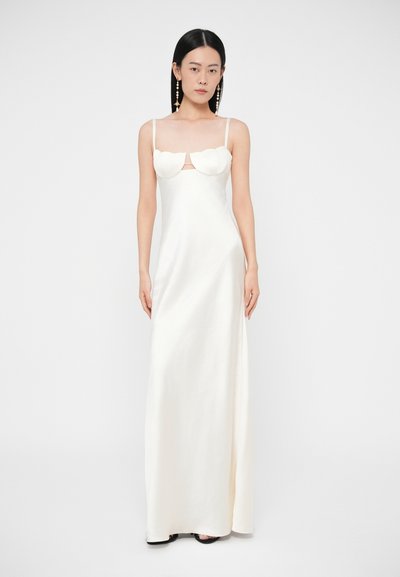 Žena s dlhými čiernymi vlasmi v dlhých bielych saténových šatách s vlnitým výstrihom a tenkými ramienkami, stojaca na jednoduchom bielom pozadí.