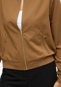 Veste brune zippée avec des zippers dorés sur un haut blanc, associée à un pantalon noir, montrant le bas du ventre et le bras droit.