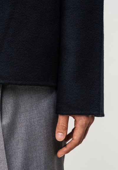 Δεξί χέρι ατόμου που φοράει σκούρο μπλε μάλλινο παλτό και γκρι παντελόνι πάνω σε ουδέτερο φόντο.
