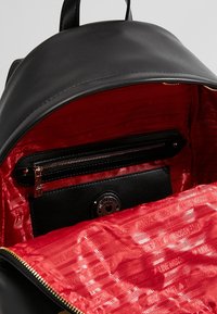 Černý batoh se zipovým uzávěrem; červená podšívka má vzorovaný design. Obsahuje malou černou kapsu na zip.