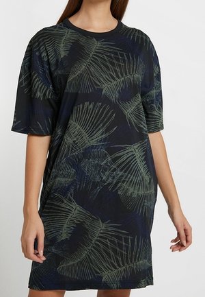 Kobieta ubrana w luźną, czarną sukienkę z krótkim rękawem, sięgającą kolan, ozdobioną zielonymi, abstrakcyjnymi wzorami liści.