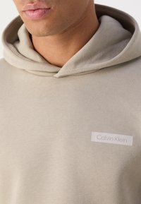 Svijetlosmeđa dukserica od mekog materijala, s istaknutom oznakom Calvin Klein ušivenom sprijeda.