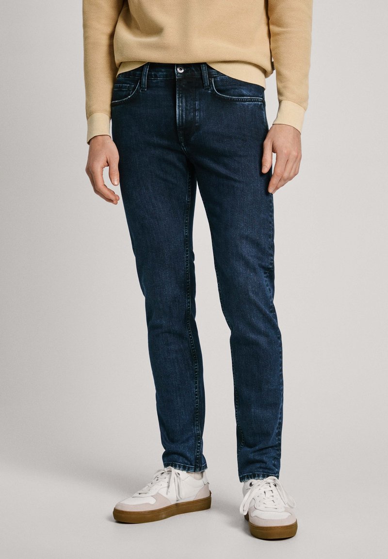 Homme portant un jean slim bleu marine, un haut à manches longues beige et des baskets blanches avec des semelles marron, debout devant un fond uni.