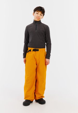 Zwarte fleece pullover met halve ritssluiting en logo, gecombineerd met feloranje sneeuwbroeken met een riem en zijzakken, gedragen met zwarte laarzen.