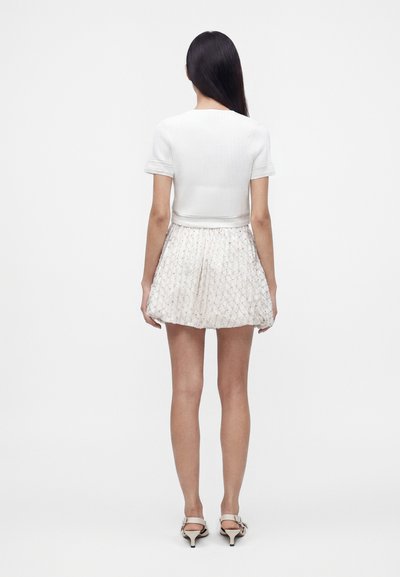 Femme aux longs cheveux foncés portant un haut blanc côtelé à manches courtes, une jupe blanche texturée et des sandales blanches à talons, debout de dos.
