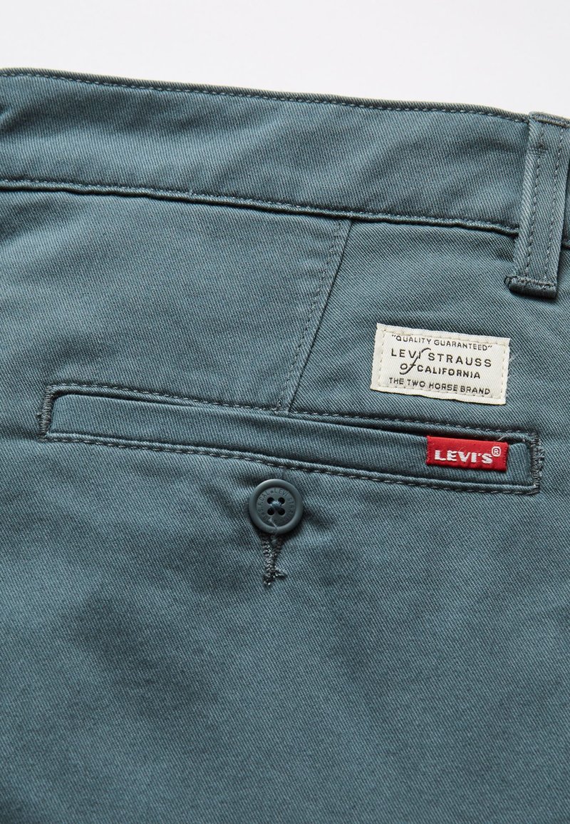 Pantalons en coton bleu-vert clair avec un bouton et une poche arrière, arborant une étiquette de marque blanche et un onglet rouge Levi's. Texture lisse.