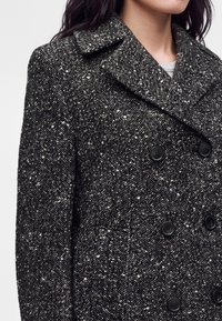 Femme portant un manteau en laine gris foncé à double boutonnage avec des boutons noirs et un motif en chevrons avec des taches blanches, col visible.