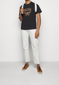 Camiseta de algodón negra con un logo naranja, pantalones blancos texturizados y zapatillas marrones con cordones negros; mochila blanca ajustable con acentos.