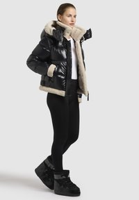 Zwarte pufferjack met beige faux shearling voering, ritszakken en verstelbare capuchon, gecombineerd met zwarte leggings en boots.