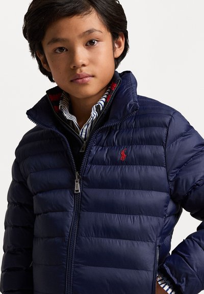 Marineblå pufferjakke med horisontalt quiltet mønster, høy krage, glidelås og rød logoakcent. Brukt over en stripete skjorte.