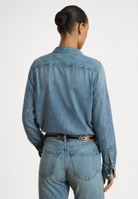 Žena s vlasmi zviazanými do drdolu, oblečená v svetlomodrej rifľovej košeli zastrčenej do zladených džínsov s čiernym opaskom, viditeľná zozadu.