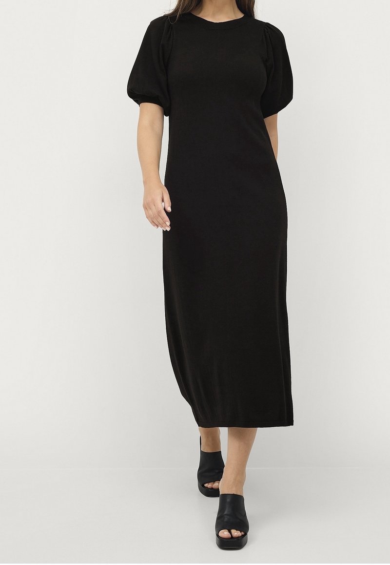 Femme portant une robe noire à manches courtes, longue jusqu'aux chevilles, avec des manches bouffantes et des chaussures ouvertes noires, debout devant un fond uni.