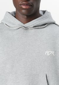 Pelēks kapucis krekls, kas izgatavots no mīksta auduma, ar priekšējo kabatu un mazu baltu "ADR" logotipu uz krūšu. Strukturālas detaļas kapuces iekšpusē.