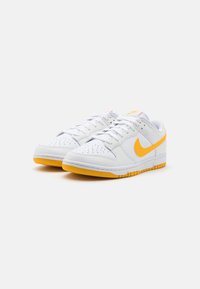 Weiße Leder-Sneaker mit gelben Akzenten, ausgestattet mit einer perforierten Zehenpartie, runden Schnürsenkeln und einer gemusterten Gummisohle. Das Nike-Logo ist sichtbar.