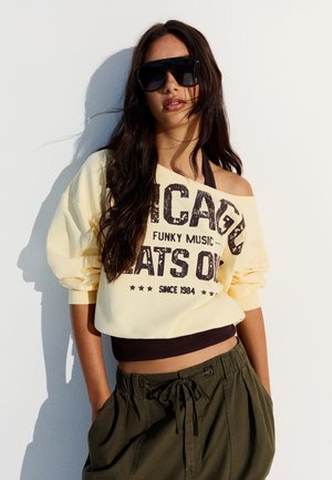 Ung kvinde med langt mørkt hår iført oversized sorte solbriller, en gul off-shoulder sweatshirt med tekst og olivengrønne bukser.