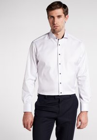 Camicia bianca a maniche lunghe con colletto a punte, bottoni neri e una texture liscia, abbinata a pantaloni scuri.