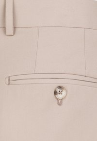 Beżowe spodnie o teksturowanej tkaninie, z małą przednią kieszonką i błyszczącym, okrągłym zapięciem na guzik.