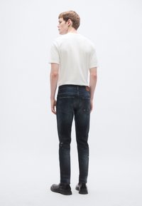 Férfi testhez álló öltözék, egyszerű fehér rövid ujjú pólóval, sötétkék szakadt farmernadrággal és fekete cipővel. Hátulnézet.