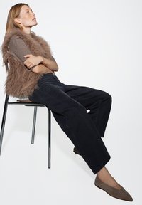 Gilet en fausse fourrure brune sur un tee-shirt gris à rayures, assorti à un pantalon large noir et des ballerines taupe, assise sur une chaise en chrome devant un fond blanc.