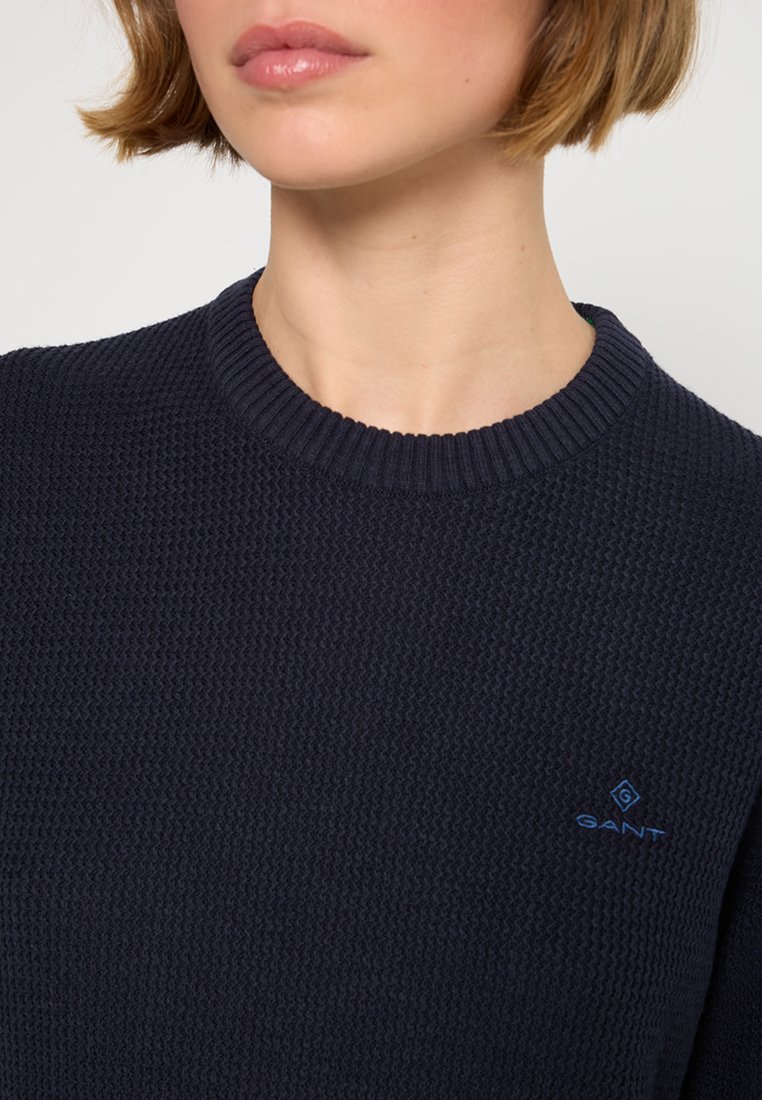 Osoba w granatowym, teksturowanym swetrze z dzianiny z prążkowanym okrągłym dekoltem oraz małym, niebieskim logo GANT na piersi.