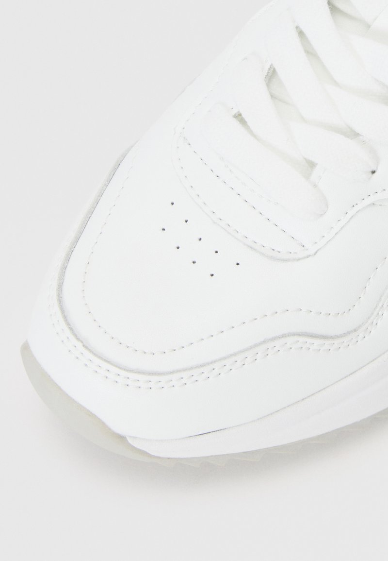 Primer plano de la parte delantera de una zapatilla blanca con cordones, perforaciones y costuras detalladas sobre un fondo claro.
