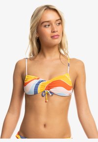 Młoda kobieta z blond włosami, nosząca kolorowy, wzorzysty biustonosz bikini z regulowanymi ramiączkami i centralnym wiązaniem, patrzy w prawo.