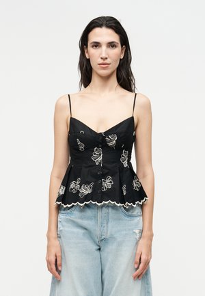 Femme aux longs cheveux foncés portant un haut noir sans manches brodé de fleurs avec boutons et un jean taille haute bleu clair.