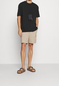 Sort t-shirt med korte ærmer, beige cargo shorts med lommer og brune sandaler. Kompakt sort pouch båret om halsen.