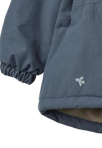 Sininen takki, jossa on kerätyt hihansuut ja pehmeä vuori sisäpuolella. Takissa on teksturoitu ulkopinta ja pieni logo alaosassa edessä.