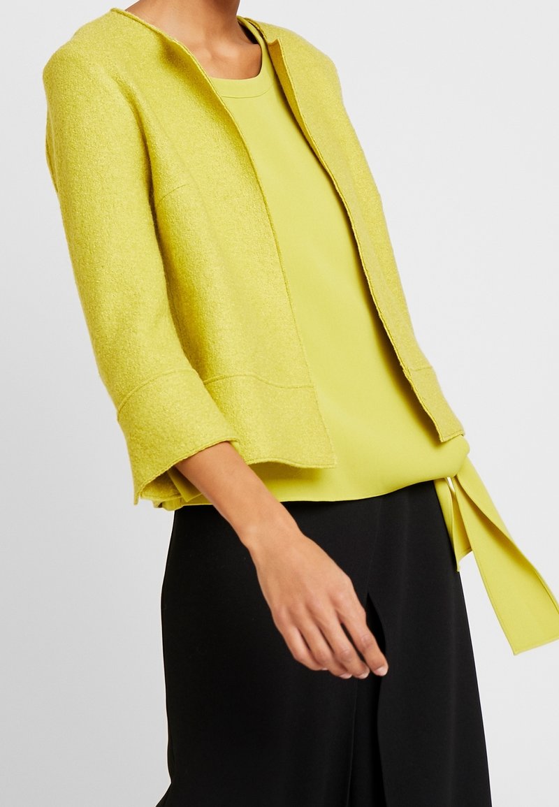 Femme portant une veste ouverte texturée vert clair sur un haut assorti avec un détail de nouage sur le côté et un pantalon large noir.
