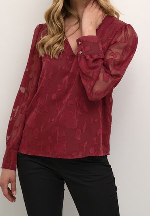 Femme portant un chemisier bordeaux transparent à manches longues avec des motifs floraux et un pantalon noir, touchant son cou d'une main.