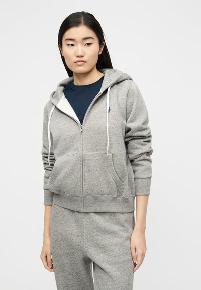 Γυναίκα που φοράει γκρι φούτερ με φερμουάρ και λευκά κορδόνια, καθώς και ταιριαστό γκρι παντελόνι φόρμας, στέκεται μπροστά σε μια απλή λευκή επιφάνεια.
