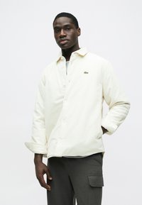 Világos krémszínű kordbársony ing gombos elejjel, mellkasi logóval és felhajtott ujjakkal. Szürke póló fölött viselve, sötét nadrággal párosítva.