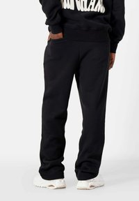Schwarze Fleece-Jogginghosen mit elastischem Bund, seitlichen Taschen, gerippten Bündchen und lockerer Passform. Kombiniert mit weißen Sportschuhen.