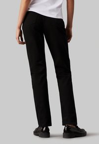 Jeans de mezclilla negra con un corte recto, que presentan bolsillos traseros y costuras visibles, combinados con zapatos negros brillantes.