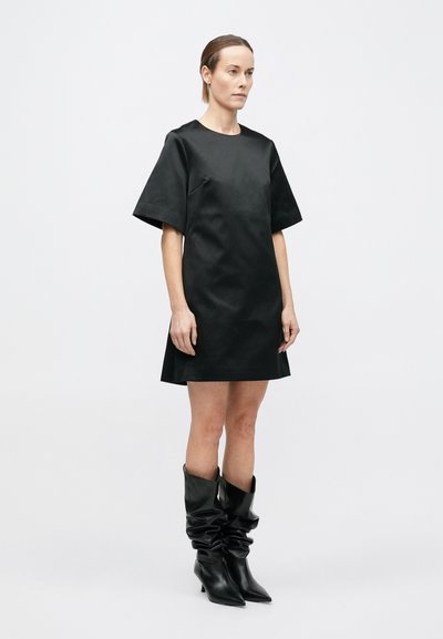 Robe noire à manches courtes avec un col arrondi et une coupe droite. Elle présente une texture lisse, associée à de grandes bottes noires décontractées.