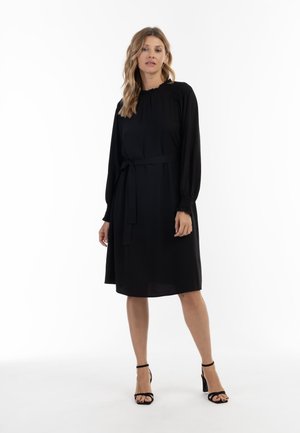 Donna con lunghi capelli biondi che indossa un vestito nero al ginocchio con maniche lunghe e sandali neri con tacco alto, in piedi su sfondo bianco.