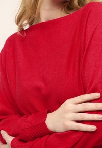 Pull en maille rouge avec un large col et des poignets côtelés, présentant une texture douce et une coupe décontractée. Une main visible repose sur le tissu.