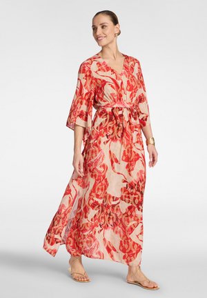 Vrouw die een vloeiende maxi-jurk draagt met een abstract rood-beige patroon, driekwart mouwen, een gouden manchette en beige sandalen, glimlachend en wandelend.