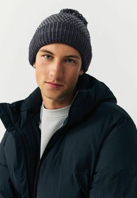 Knit beanie in shades of dark gray with a pom-pom on top; worn with a dark puffer jacket and light gray t-shirt. Smooth texture.